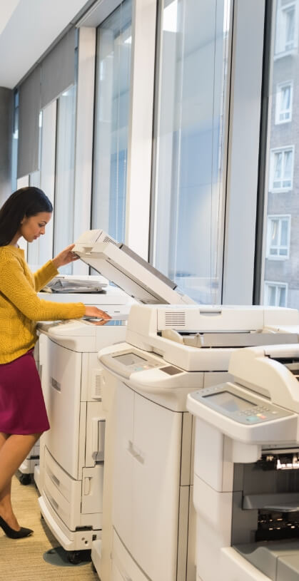 Eine junge Frau bedient einen Multifunktionsdrucker. Dieser steht in einem Gang neben zahlreichen weiteren Druckern und Scannern.