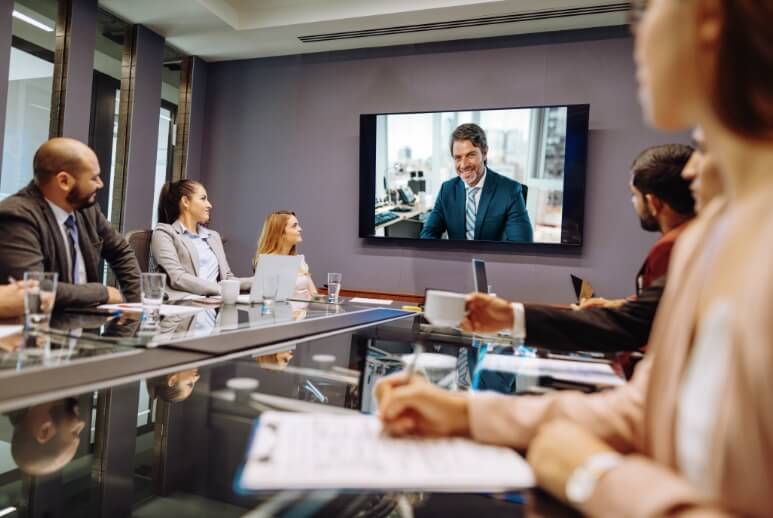 Eine Büroszene bei einem Online-Meeting. Mehrere Mitarbeiter sprechen mit einem Kollegen, der über Video-Stream zugeschaltet ist.